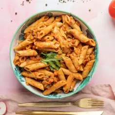 Miseczka pełna makaronu penne z sosem pomidorowym i soczewicą. Obok pomidory, rzodkiewki i bazylia. Idealna porcja, by ustalić, ile makaronu penne na osobę.