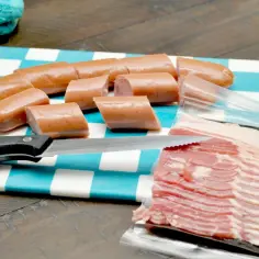 Kiełbaski pokrojone na desce w kratkę, obok plastry boczku. Zastanawiasz się, ile gram mięsa na osobę?