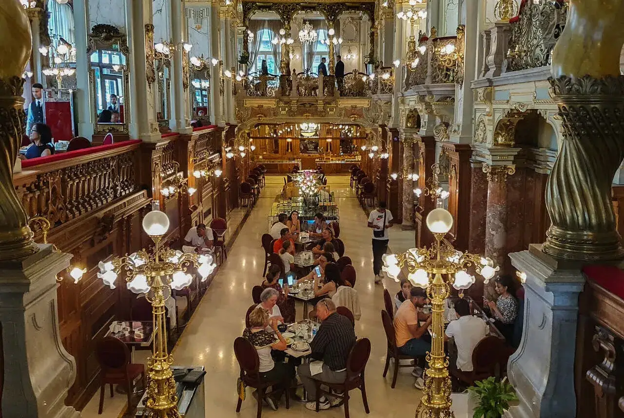 Elegancka kawiarnia w Budapeszcie, gdzie zjeść można w historycznym wnętrzu. Ludzie siedzą przy stolikach, delektując się posiłkami.