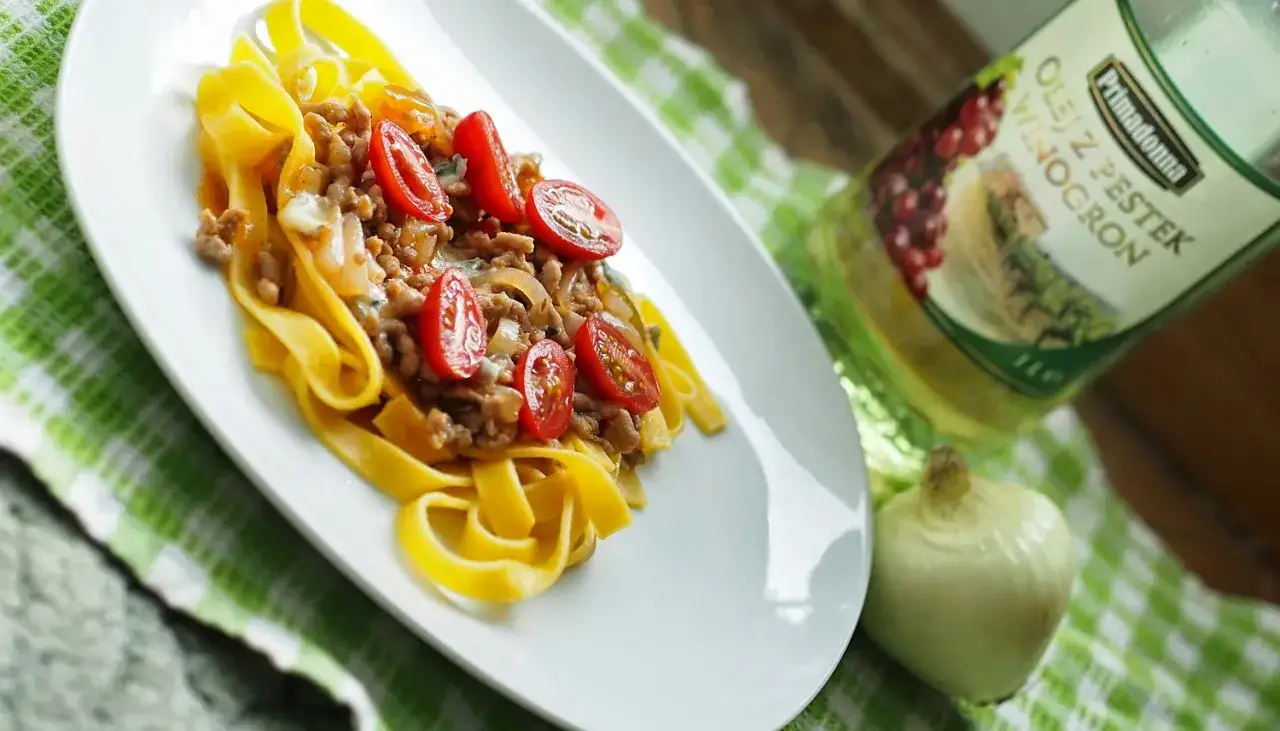 Talerz tagliatelle z mielonym mięsem i pomidorkami. Obok butelka oleju z pestek winogron i cebula. Idealne składniki, by dowiedzieć się, jak zrobić spaghetti ze słoika.