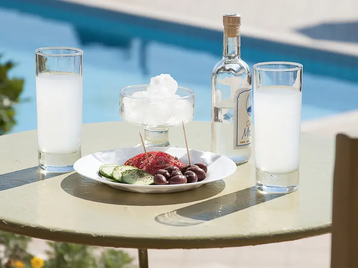 Butelka ouzo, szklanki z napojem, talerz z oliwkami i ogórkiem, lód na stole przy basenie.