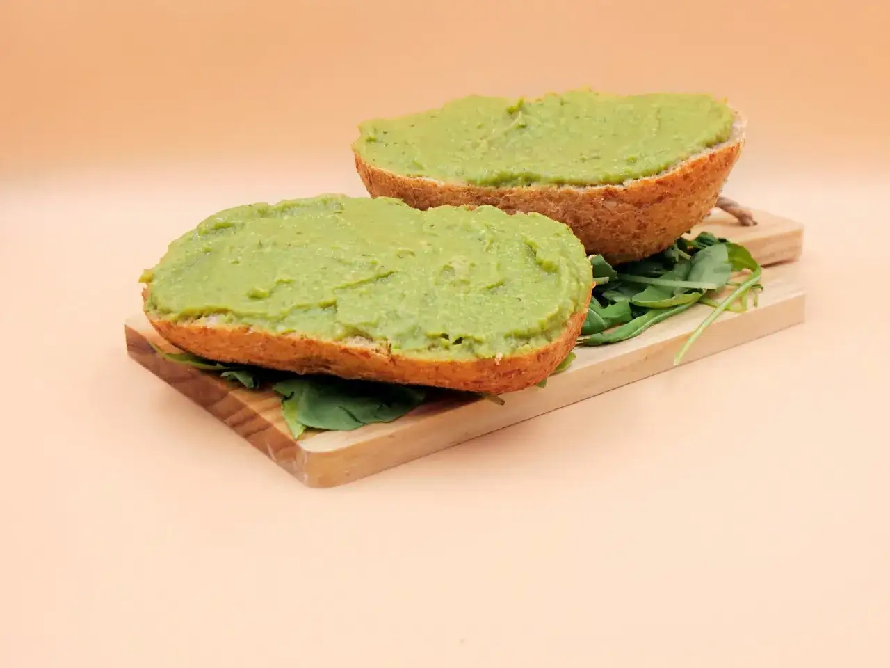 Dwie połówki bułki z zielonym, kremowym guacamole i listkami rukoli na desce. Czy guacamole jest zdrowe? Tak, to pyszna i zdrowa przekąska!