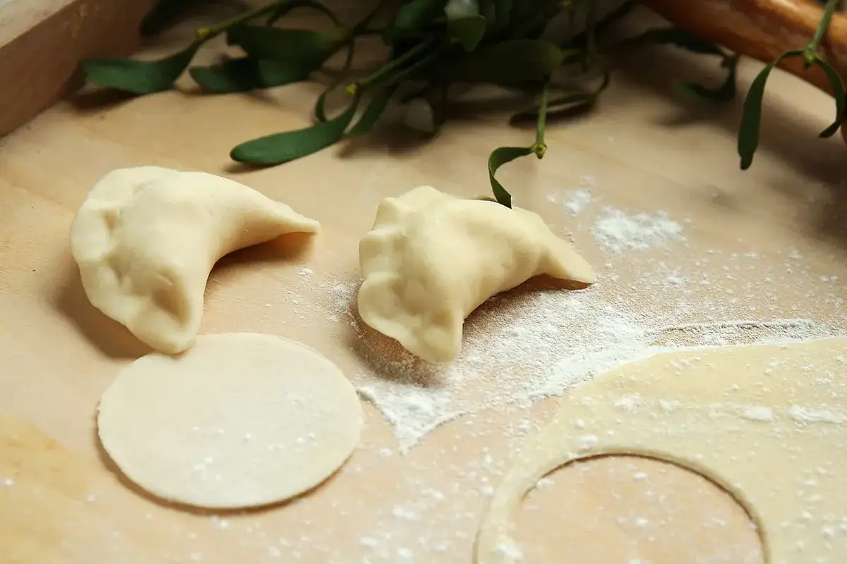 Surowe pierogi leżą na drewnianej desce, obok kółka ciasta i gałązki jemioły. Idealna mąka na pierogi to sekret pysznego dania.