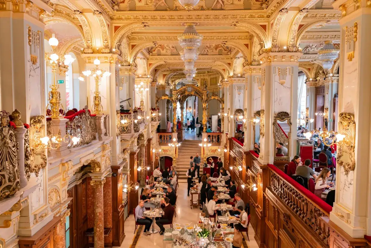 Elegancka kawiarnia w Budapeszcie, gdzie zjeść można w otoczeniu złota i kryształowych żyrandoli. Wnętrze pełne ludzi delektujących się posiłkiem.